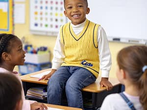 A smiling boy sitting on a desk in a classroom with classmates.