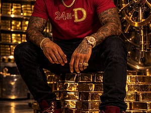Man with tattoos sitting on shiny gold barrels in stylish attire.