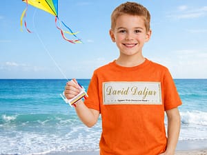 A smiling boy in an orange shirt flies a colorful kite on the beach.